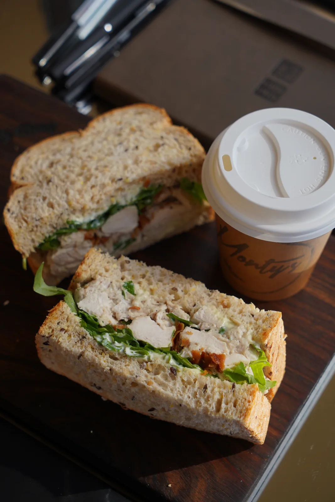 Two halves of a chicken sandwich on seeded bread next to a Monty's takeaway coffee cup on a timber board