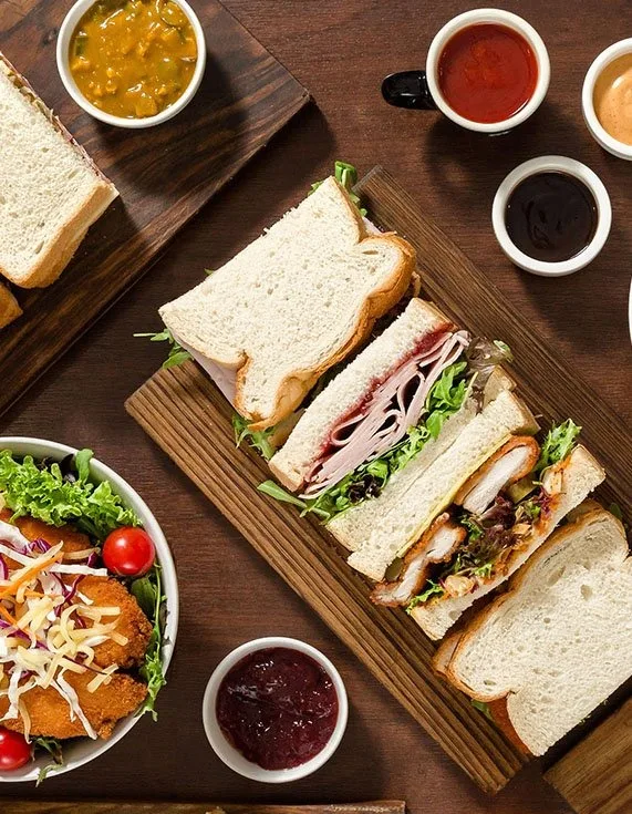 Sandwiches on white bread cut in halves on wooden boards, with a salad bowl and small pots of sauce